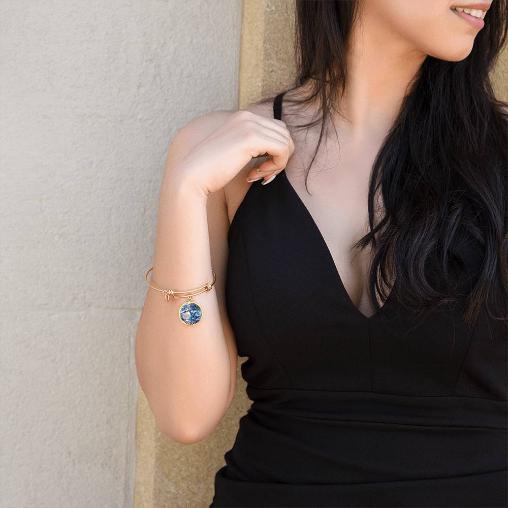 A woman with long dark hair is wearing a black sleeveless V-neck dress and gold bangle bracelets. One bracelet features the Aquarius Zodiac Bangle Bracelet | Personalized Zodiac Bangle by ShineOn Fulfillment. She is standing against a neutral-colored wall, with her right hand raised to her shoulder and smiling slightly.