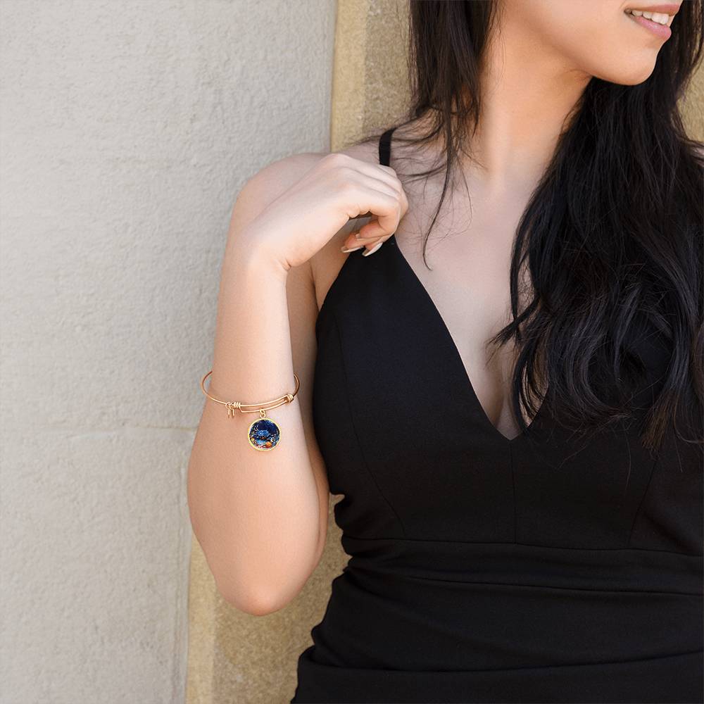A woman with long black hair wearing a sleeveless, v-neck black dress leans against a beige wall. She has a ShineOn Fulfillment Cancer Zodiac Bangle Bracelet With Optional Personalization on her left wrist that features a large blue and gold circular charm. The woman is looking slightly away from the camera.