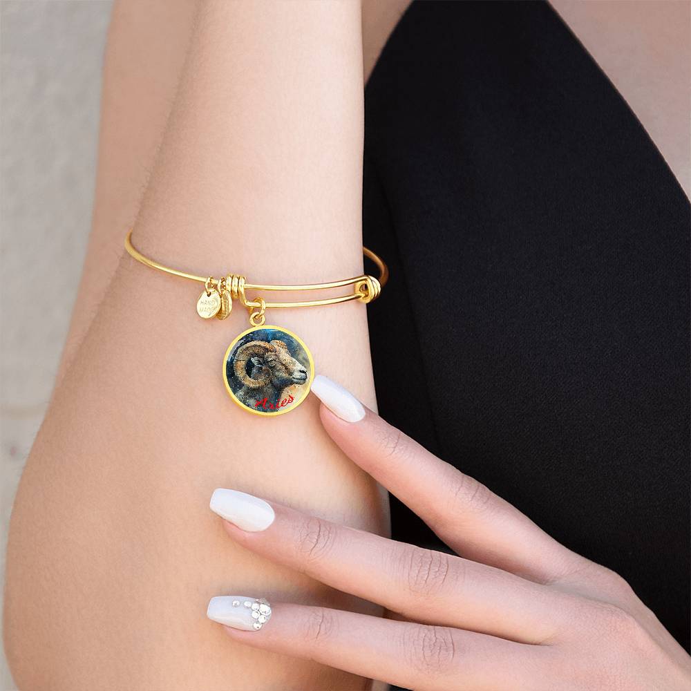 A person with a black sleeveless top is wearing a gold Aries Zodiac Bangle Bracelet With Optional Personalization from ShineOn Fulfillment. The central charm features a detailed depiction of a ram. The high-quality surgical steel bracelet includes personalized jewelry options with custom engraving. The person has manicured nails painted in light colors with decorative accents.