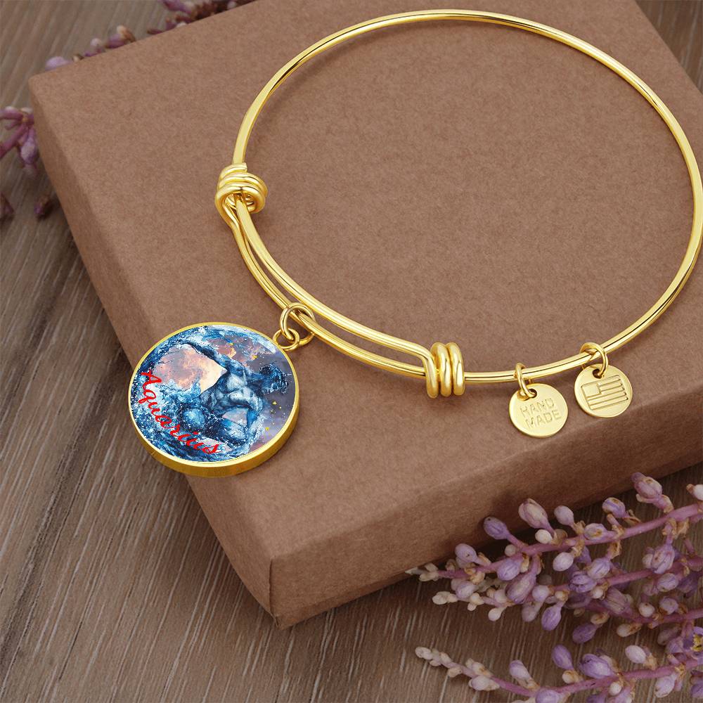 An Aquarius Zodiac Bangle Bracelet | Personalized Zodiac Bangle with two small round charms, one reading "HANDMADE" and the other with a striped design, sits atop a brown cardboard box. A larger round pendant with a blue and red abstract design hangs from this piece of personalized jewelry. Purple flowers are placed around the box, all crafted by ShineOn Fulfillment.