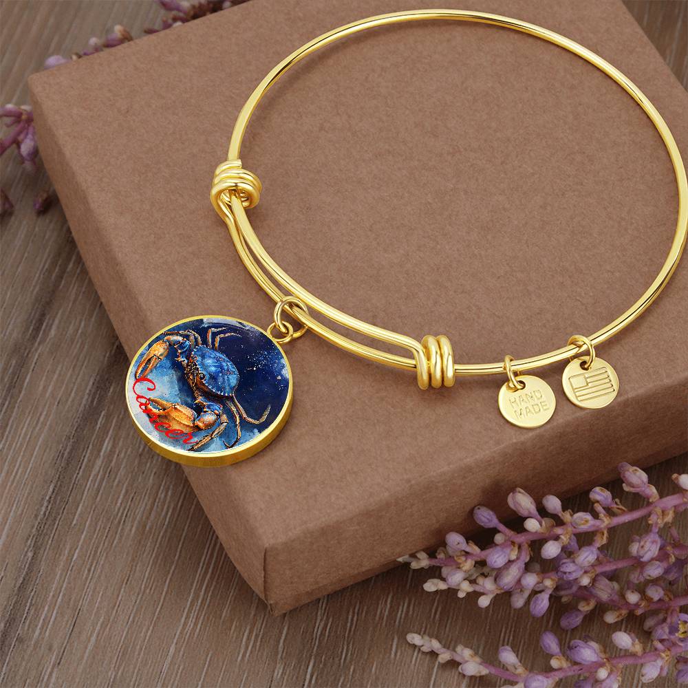 A Cancer Zodiac Bangle Bracelet With Optional Personalization by ShineOn Fulfillment is displayed on a brown box with small lavender flowers around it. The personalized jewelry features a round pendant with a detailed, colorful crab illustration and two smaller charms: one reads "HANDMADE" and the other has a striped design.