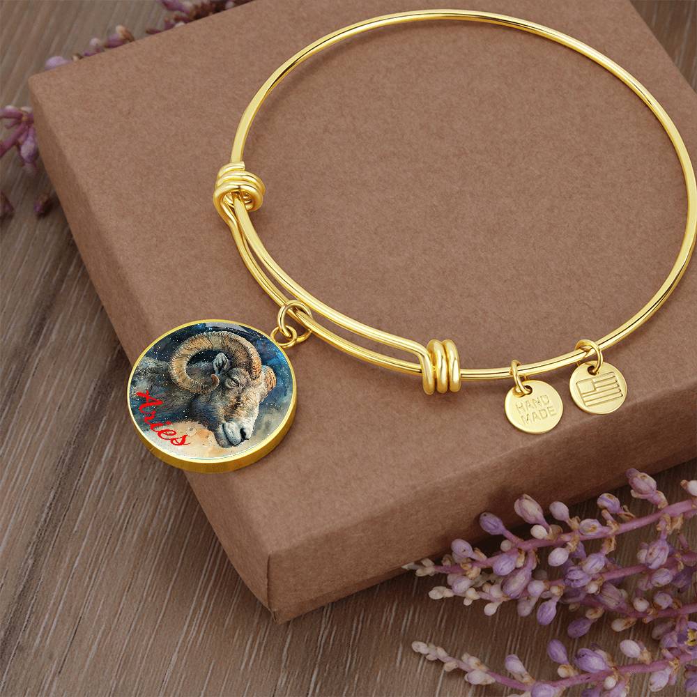 A gold Aries Zodiac Bangle Bracelet With Optional Personalization by ShineOn Fulfillment sits atop a brown cardboard box, adorned with dried lavender. The personalized jewelry features three charms: a round one with a ram illustration and the word "Aries" in red, a "HAND MADE" tag, and a circular tag with horizontal lines.