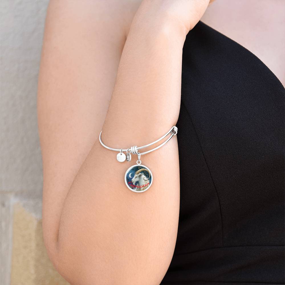 A person wearing a black sleeveless top showcases a ShineOn Fulfillment Capricorn Zodiac Bangle Bracelet With Optional Personalization on their arm. The bracelet features two charms: a small circular disc and a larger charm with a colorful image. The arm is bent with the hand resting near the shoulder, highlighting this piece of personalized jewelry.