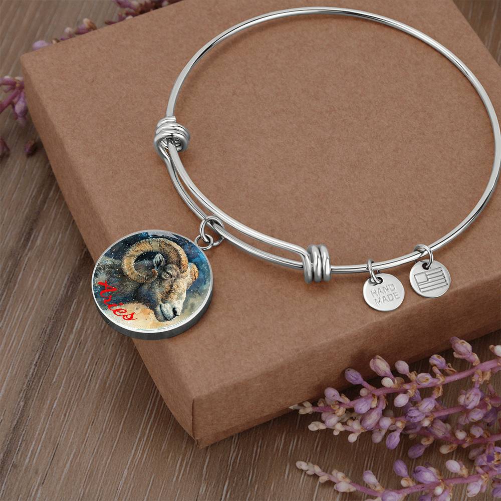 The Aries Zodiac Bangle Bracelet With Optional Personalization from ShineOn Fulfillment features a silver bangle bracelet with three charms: a round pendant featuring an illustration of a ram with "Aries" written in red, a small heart-shaped charm with "HAND MADE" engraved, and a circular charm with an abstract design. The personalized jewelry rests on a brown box, surrounded by purple flowers.