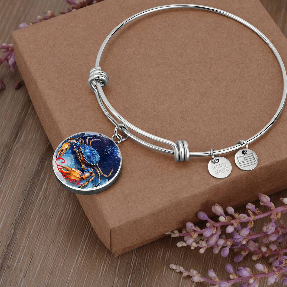 A Cancer Zodiac Bangle Bracelet With Optional Personalization by ShineOn Fulfillment, featuring a round pendant with a painted blue crab and two small silver charms, one stamped with "HAND MADE" and the other with an abstract design, crafted from surgical steel, is displayed on a brown cardboard box surrounded by small purple flowers.