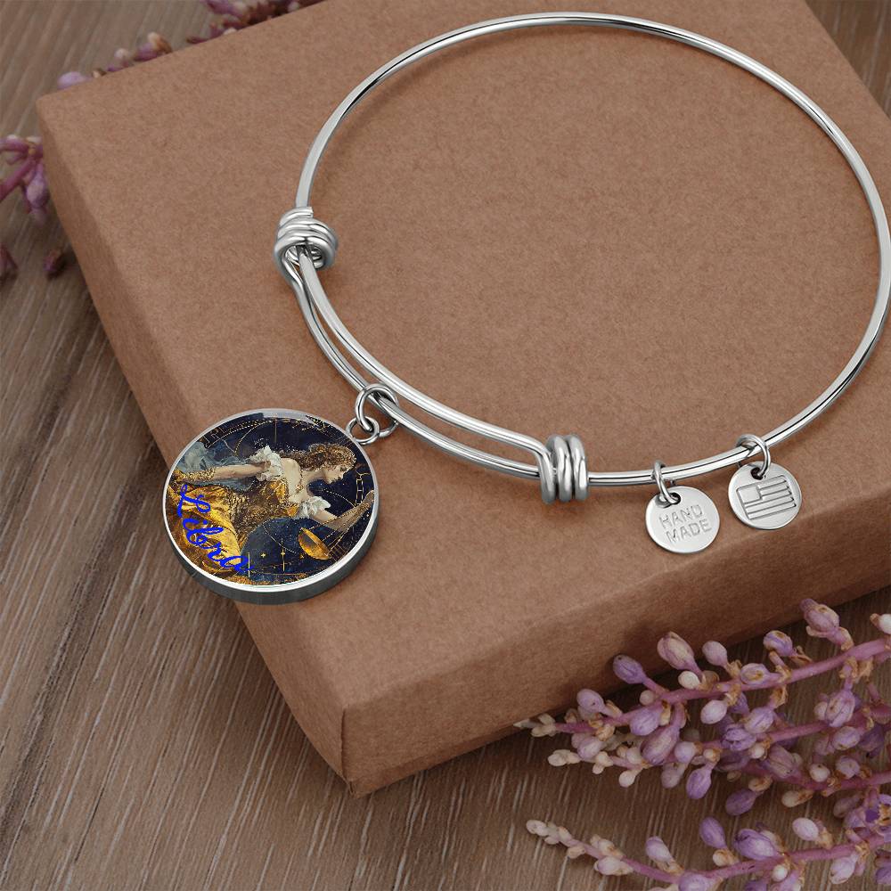 A Libra Zodiac Bangle Bracelet With Optional Personalization from ShineOn Fulfillment with a round charm boasting a colorful, intricate design rests on a brown gift box surrounded by small purple flowers. The personalized jewelry includes two smaller charms, one inscribed with "HANDMADE" and the other with parallel lines, adding an elegant touch.
