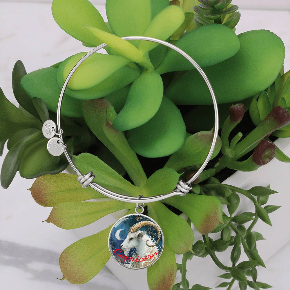 A silver bangle bracelet with an 18k gold finish featuring a Capricorn charm with a goat illustration is displayed against a backdrop of various green succulents in a white pot. The charm has an image of a goat with the text "Capricorn" in red cursive. Introducing the Capricorn Zodiac Bangle Bracelet With Optional Personalization by ShineOn Fulfillment, this unique piece combines elegance and astrological significance.